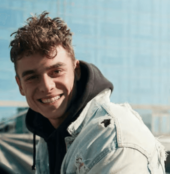Smiling young man with curly hair wearing a distressed denim jacket against a blurred background.